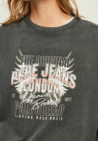 Sudadera gris oscuro con un estampado gráfico que incluye texto y un diseño de alas en blanco, rosa y verde. Cuello redondo de canalé y textura suave.