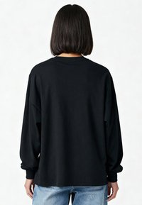 Persoon met steil, donker haar op schouderlengte, gekleed in een loszittend zwart shirt met lange mouwen en lichtblauwe spijkerbroek, van achteren gezien tegen een witte achtergrond.