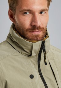 Bež jakna s visokim ovratnikom, zatvaračem na munju i crnim hardverom. Sadrži teksturiranu tkaninu i naglasak logoa na povlačenju zatvarača.