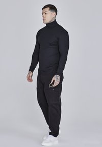 Black long-sleeve turtleneck top, fitted design; paired with black cargo trousers featuring pockets and a gold insignia, white sneakers.