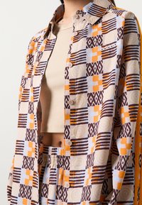 Skjorta med knappar i ett färgglatt geometriskt mönster i brunt, orange, blått och gult, kombinerad med en beige ribbad topp och matchande byxor.