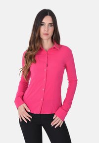 Camicia aderente a maniche lunghe di un brillante colore rosa, realizzata in tessuto liscio con abbottonatura frontale e colletto classico. Silhouette sagomata, abbinata a pantaloni neri.