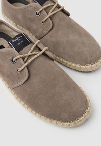 Zapatos espadrilles de ante marrón con un acabado texturizado y cordones de cuerda. Cuenta con una suela de yute trenzado y una etiqueta de marca negra cerca de los cordones.