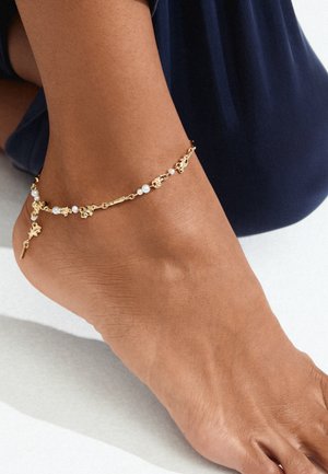 Zlatna anklet s bijelim biserima, složenim detaljima i malim privjescima; nosi se na goloj nozi na neutralnoj pozadini.