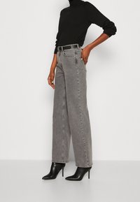 Graue Denim-Hosen mit weitem Bein und hoher Taille, mit Fronttaschen und sichtbaren Nähten, kombiniert mit einem schwarzen Rollkragenpullover und schwarzen Stilettos.