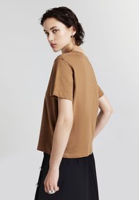 Maglietta a maniche corte marrone in taglio comodo, realizzata in tessuto testurizzato. Presenta un collo a giro e orlo dritto. Indossata con una gonna nera.