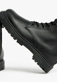 Des bottes en cuir noir présentent une surface lisse, un bout rond, un design solide à lacets et une semelle en caoutchouc texturée pour une meilleure adhérence.