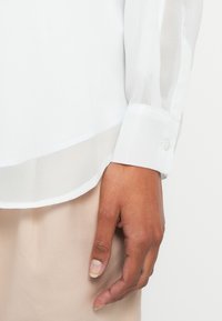 White blouse with sheer fabric, featuring a rounded hem, buttoned cuff, and a smooth texture. Hand positioned near the blouse's edge.