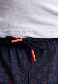 Námořnické plavky s červeným vzorem palem, s páskem na stažení s oranžovými konci. Tričko nahoře je bílé z bavlny.