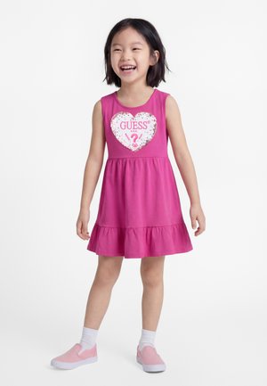 Jeune fille souriante portant une robe rose sans manches avec un logo en forme de cœur "GUESS U.S.A." et des chaussures roses sans lacets avec des chaussettes blanches.