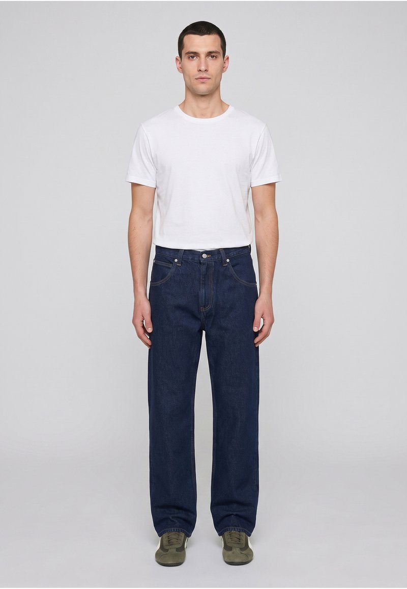 Homme debout face à l’avant, portant un t-shirt blanc uni, un jean bleu foncé et des baskets vertes sur un fond uni gris clair.