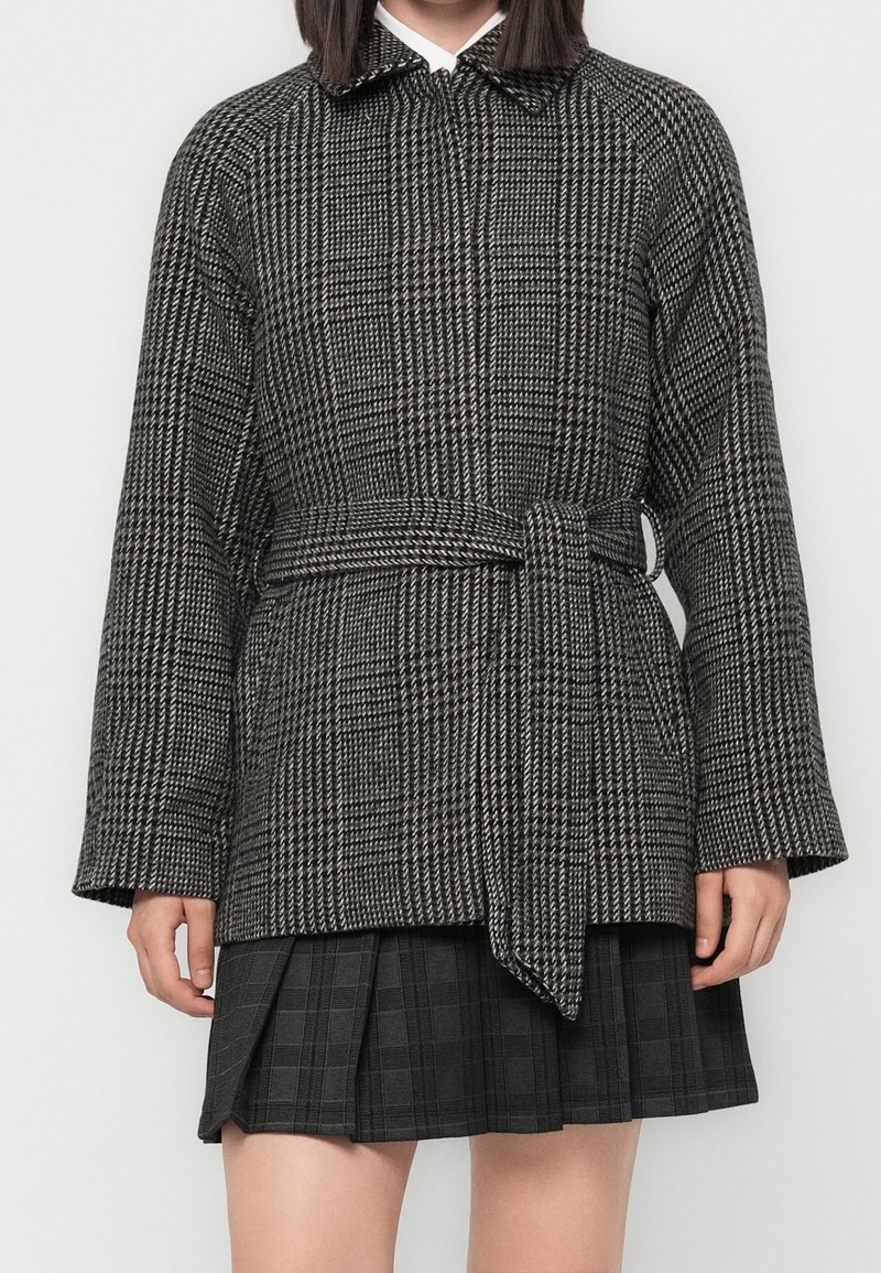 Femme portant une veste pied-de-poule noire et blanche ceinturée sur une jupe plissée sombre, debout devant un fond clair uni.