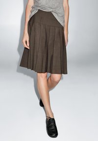 Brown pleated skirt with a smooth texture and asymmetrical waistband, paired with black lace-up shoes. Light gray top partially tucked.