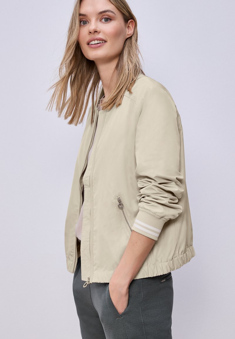 Mujer de cabello rubio que lleva una cazadora bomber beige claro con mangas fruncidas y bolsillos laterales con cremallera, de pie con una mano en el bolsillo del pantalón gris.