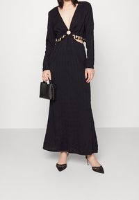 Robe noire à manches longues avec un décolleté plongeant en V, un détail noué à la taille et un accent circulaire. Le mannequin tient un petit sac noir et porte des talons pointus.