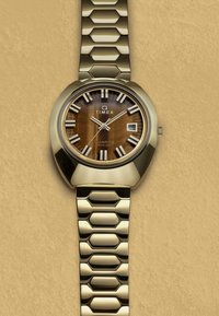 Guldtonet kvartsur med et rundt urkasse, brun trætekstureret urskive, hvide time markører og metalarmbånd med sekskantet mønster. Datovindue til stede.