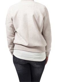 Felpa beige chiaro con una texture liscia, collo rotondo e polsini a coste, indossata sopra una maglietta bianca. La vista posteriore mostra una vestibilità comoda.