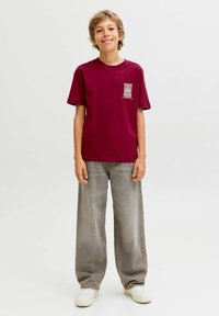 Tumši sarkans kokvilnas t-krekls ar īsām piedurknēm, uz kura kreisās krūtis ir neliela grafika. Valkāts ar brīvām pelēkām džinsām un baltajiem sporta apaviem.