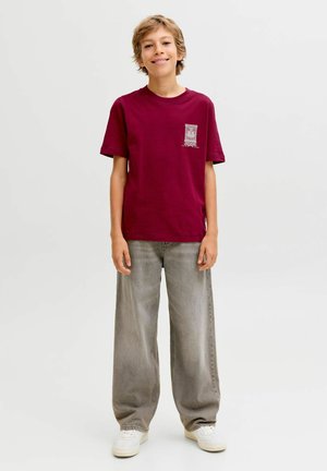 Camiseta de algodón en color marrón con mangas cortas, que presenta un pequeño diseño gráfico en el lado izquierdo del pecho. Se lleva con pantalones de mezclilla grises holgados y zapatillas blancas.