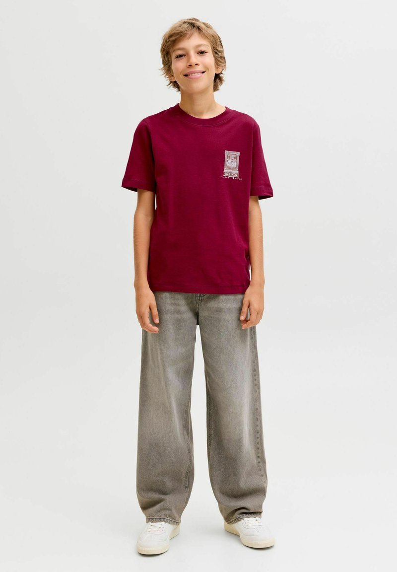 Tumši sarkans kokvilnas t-krekls ar īsām piedurknēm, uz kura kreisās krūtis ir neliela grafika. Valkāts ar brīvām pelēkām džinsām un baltajiem sporta apaviem.