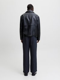 Άτομο με κοντά μαλλιά που φοράει μαύρο δερμάτινο μπουφάν, μπλε σκούρο παντελόνι με ρίγες και φαρδιές παντελόνες, και μαύρα παπούτσια, στέκεται με την πλάτη σε απλό φόντο.