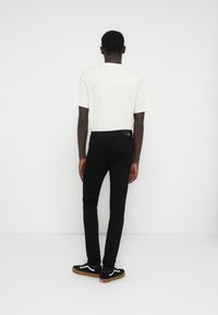 Vaqueros negros de corte slim con bolsillos traseros, combinados con una camiseta de color claro. Se llevan con zapatillas negras que tienen un logo blanco y suela marrón claro.