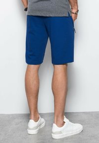 Shorts in maglia blu con una vestibilità comoda, dotati di tasche laterali e orlo dritto. Indossati con sneaker bianche su una superficie grigia.