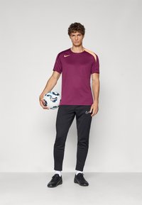 T-shirt atlético roxo com detalhes laranja, combinado com calças pretas ajustadas. O modelo segura uma bola de futebol estampada e usa sapatos pretos.