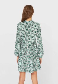 Vestito floreale verde con lunghi maniche a sbuffo, vita aderente e gonna svasata. Il tessuto presenta un motivo di margherite bianche su uno sfondo scuro.