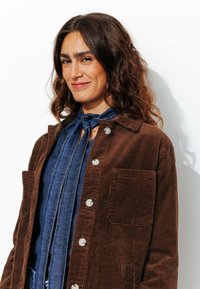 Veste en velours côtelé marron avec fermeture à bouton, deux poches avant, et une chemise en denim en dessous avec un détail de cravate nouée.