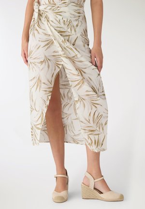 Donna che indossa una gonna avvolgente beige e bianca con motivo a foglie e espadrillas beige con zeppa e cinturini alla caviglia su sfondo bianco.