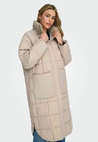 Beige vadderad dunjacka med hög krage, utrustad med två stora framfickor, dragkedja och sidospjäll för rörelse.