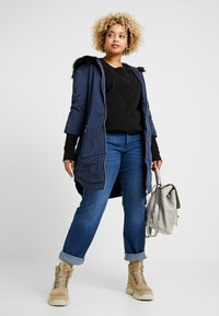 Parka marina con capucha de piel sintética, suéter negro bordado, jeans azules, botas beige con cordones y mochila gris. Las características clave incluyen bolsillos y texturas.