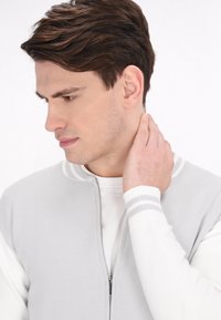 Cárdigan de cremallera gris claro con mangas largas blancas. Presenta un cuello acanalado y puños a rayas. La modelo se toca el cuello, mirando ligeramente hacia la izquierda.
