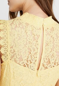 Žlutý krajkový top s jemnými květinovými vzory, vysokým výstřihem a zapínáním na knoflíky na zadní straně. Lehký s průsvitným designem.