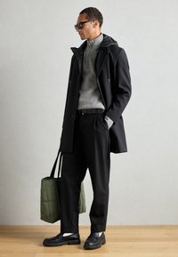 Svart frakk med hette, grå strikkegenser, svarte bukser, svarte slip-on sko og en grønn quiltet tote bag. Glatt tekstur og moderne design.