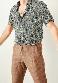 Kurzärmliges Hemd mit einem grün-blauen Tropfenmuster auf hellen Hintergrund, kombiniert mit braunen gestreiften Jogginghosen mit Kordelzug.