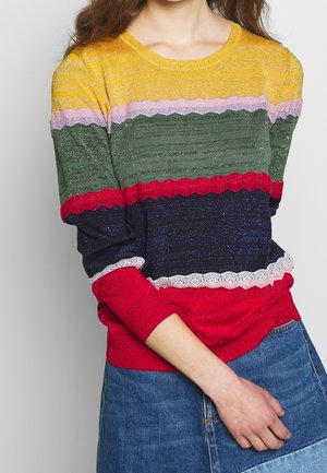 Personne portant un pull multicolore à rayures horizontales jaune, violet, vert, rouge, bleu marine et blanc, associé à une jupe en jean bleue.