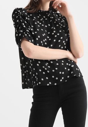 Femme portant un chemisier noir à manches courtes avec des imprimés de fleurs blanches et des épaules froncées, associé à un pantalon noir.