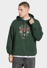 Zielona bluza z kapturem z kieszenią z przodu, ozdobiona graficznym nadrukiem w czerwonym i białym kolorze. Wykonana z bawełny, ma luźny krój i kaptur z regulowanym sznurkiem.