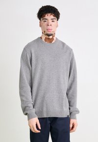 Gray knit sweater with a round neckline, ribbed cuffs and hem, textured fabric, and a tag at the bottom, worn over navy trousers.