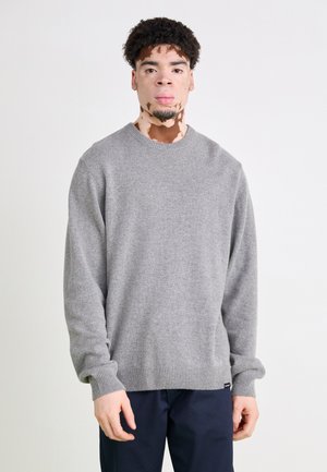 Gray knit sweater with a round neckline, ribbed cuffs and hem, textured fabric, and a tag at the bottom, worn over navy trousers.
