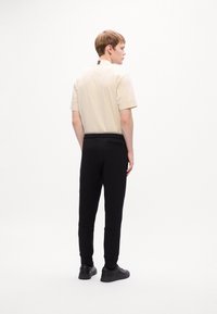 Jonge man staat, draagt een beige overhemd met korte mouwen dat in zwarte broek is gestoken, met zwarte sneakers, met zijn rug naar de camera gericht tegen een effen witte achtergrond.