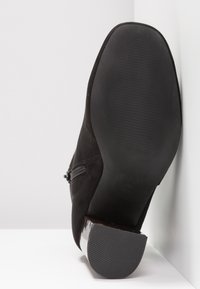 Bota de tobillo de ante negro con un tacón negro brillante, suela de goma texturizada y un detalle de cremallera en el exterior. Superficie lisa y punta redondeada.