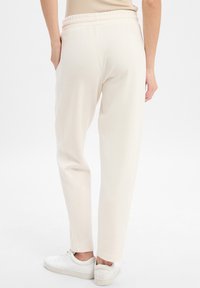 Pantalon de jogging beige clair avec une coupe décontractée, taille élastique et poches latérales, associé à des baskets blanches sur un fond uni.