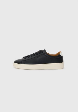Tenis de couro preto com uma textura suave, sola de borracha bege, design de atar e um destaque no calcanhar em contraste.