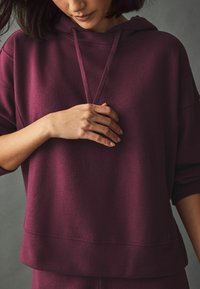 Burgundská oversized mikina vyrobená z mäkkého, textúrovaného materiálu, s kapucňou na šnúrku a spustenými ramenami, s uvoľneným strihom a rebrovaným lemom.