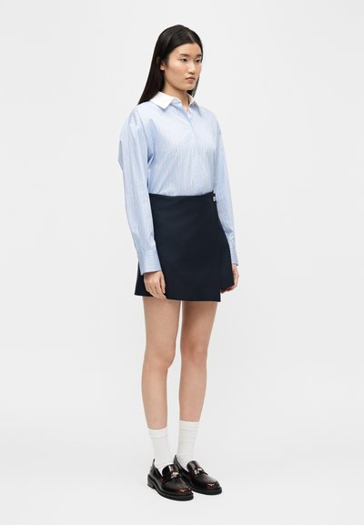 Γυναίκα που στέκεται, φορώντας ένα ανοιχτό γαλάζιο πουκάμισο με ρίγες, μίνι φούστα σε ναυτικό μπλε με κουμπιά στο πλάι, λευκές κάλτσες και μαύρα μοκασίνια, μπροστά σε απλό φόντο.