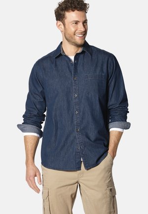 Dunkelblaues Jeanshemd mit Knopfverschluss, Brusttasche und hochgerollten Ärmeln über einer weißen Unterlage. Getragen mit khakifarbenen Cargo-Hosen.