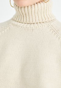 Cremfarbener gestrickter Pullover mit einem hohen, gerippten Kragen und strukturiertem Oberteil. Besitzt sichtbare Nähte und eine entspannte Passform.